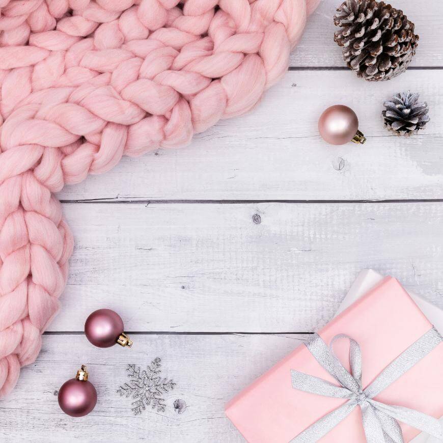 A pink chunky knit blanket, pinecones, pink baubles, a pink gift box with a silver ribbon, and a silver snowflake ornament arranged on a white wooden surface.