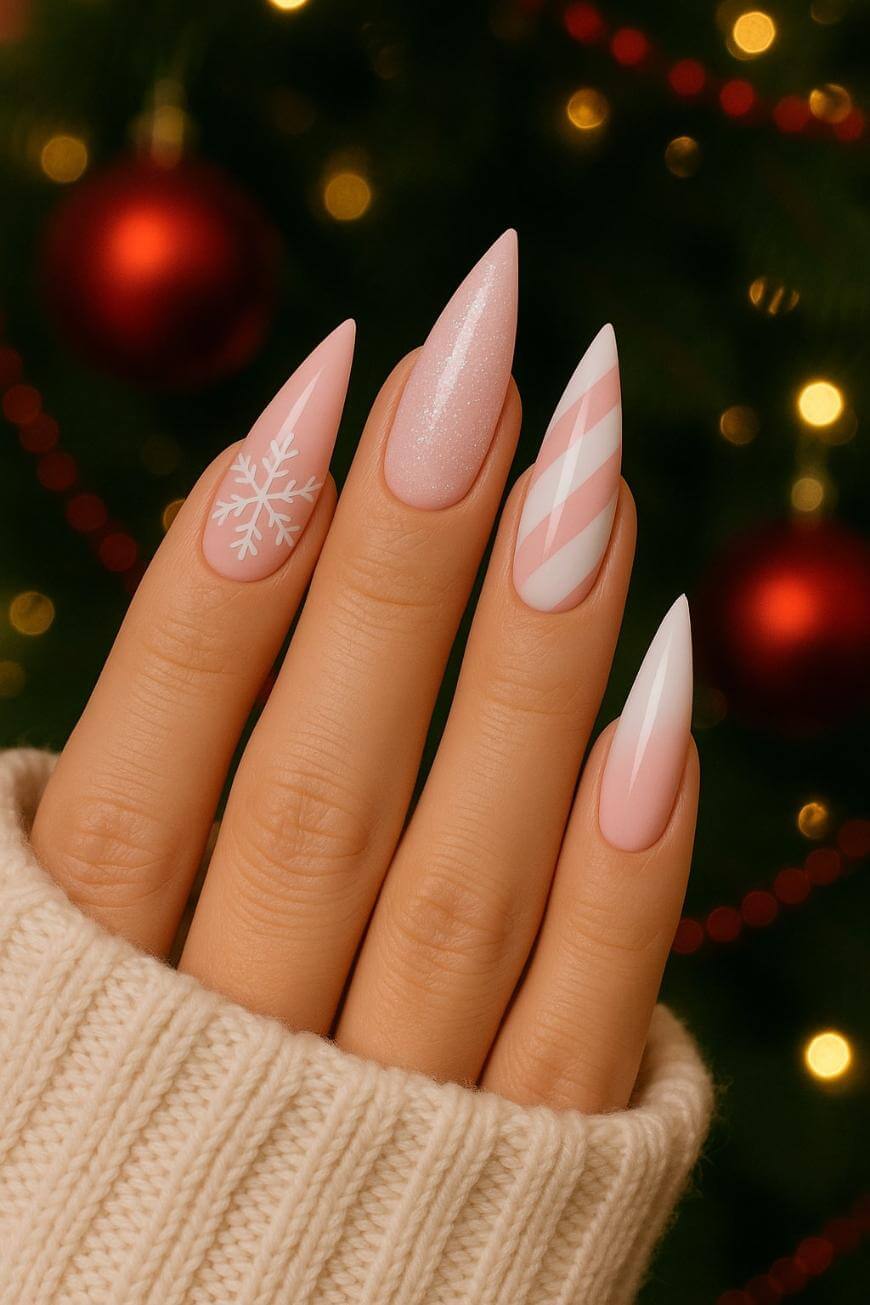 A hand with long, almond-shaped nails featuring festive nail art: a white snowflake, pink glitter, pink and white stripes, and a pink and white tip. The person is wearing a cream sweater, with a blurred Christmas tree in the background.