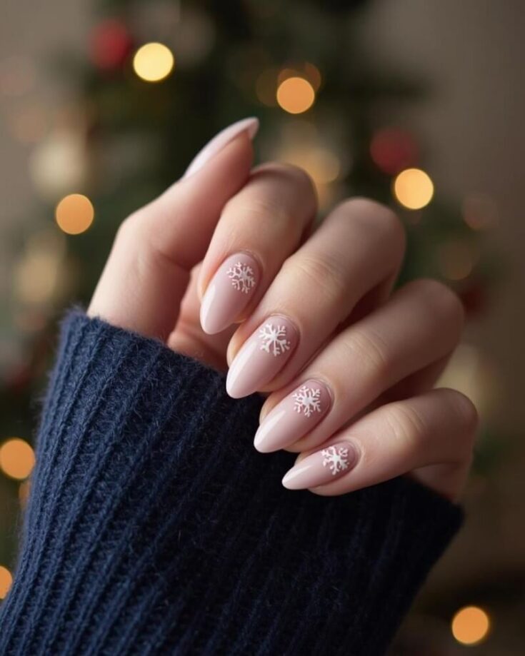 A hand with long, almond-shaped nails painted a pale pink, each nail decorated with a white snowflake design. The person is wearing a dark blue sweater, and blurred Christmas lights are in the background.