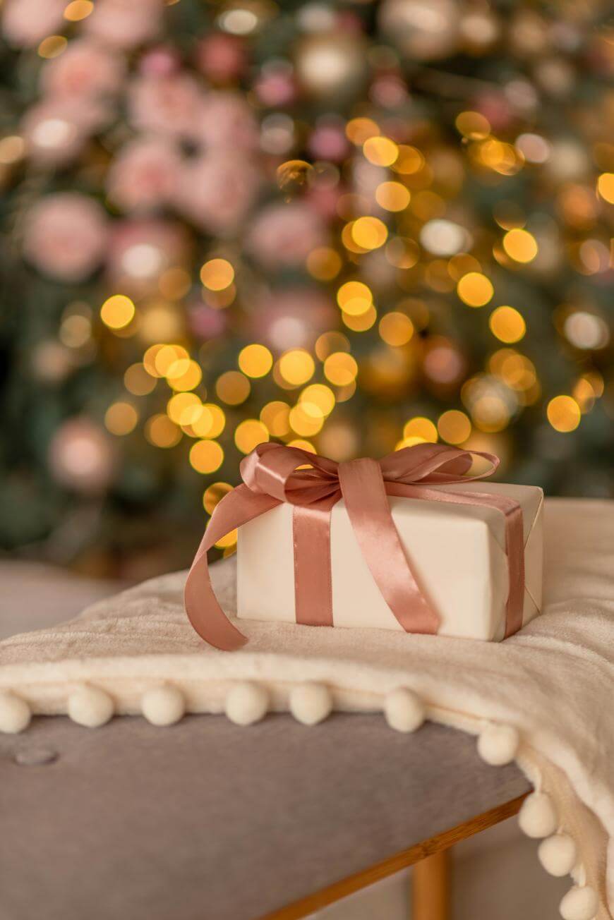 A gift wrapped in cream paper with a pink satin ribbon sits on a soft, white blanket with pom-poms. In the blurred background, warm yellow holiday lights and ornaments decorate a Christmas tree.