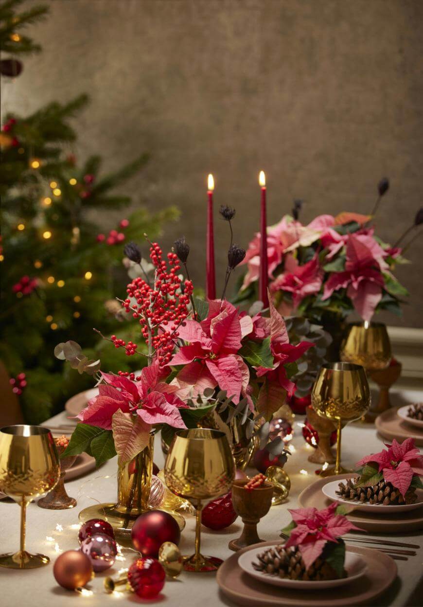 A festive holiday table is set with gold glasses, pink poinsettia centerpieces, red berries, and pinecones. Red ornaments and lit candles add a warm, elegant touch. A decorated Christmas tree is in the background.