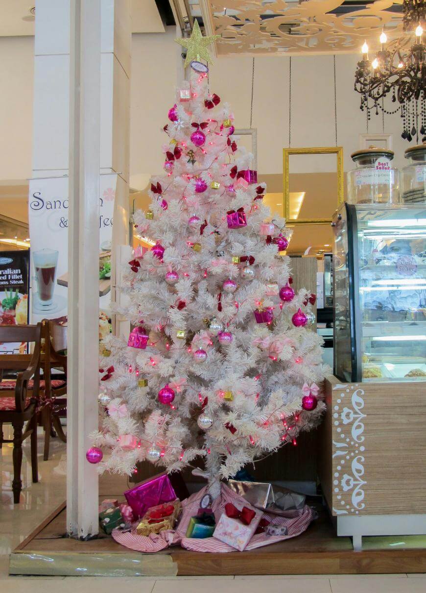 A white Christmas tree decorated with pink and gold ornaments stands indoors beside a glass display case, with wrapped gifts in pink and gold under the tree.