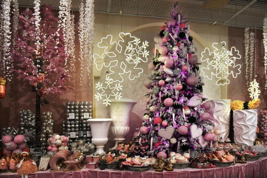 A Christmas display features a pink and purple decorated tree with heart and sphere ornaments, surrounded by festive decorations, faux pink trees, and large white vases. White snowflake lights hang in the background.
