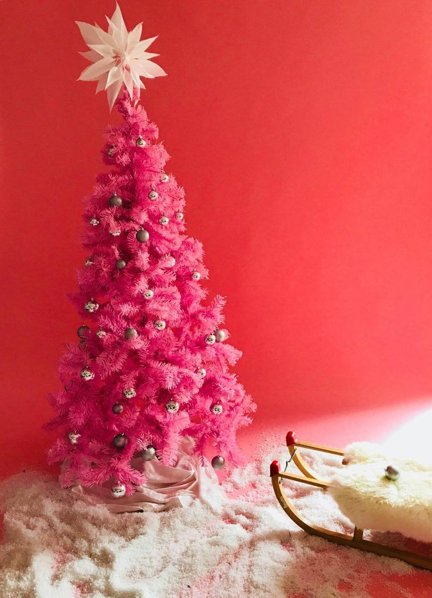 A bright pink Christmas tree decorated with silver ornaments stands on fake snow next to a vintage wooden sled with a fur cover, set against a vibrant pink background.