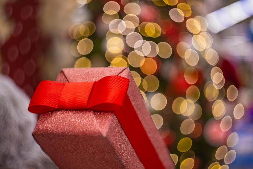 A red gift box with a bright red ribbon is in focus, with a blurred background of warm, festive lights and an out-of-focus Christmas tree.