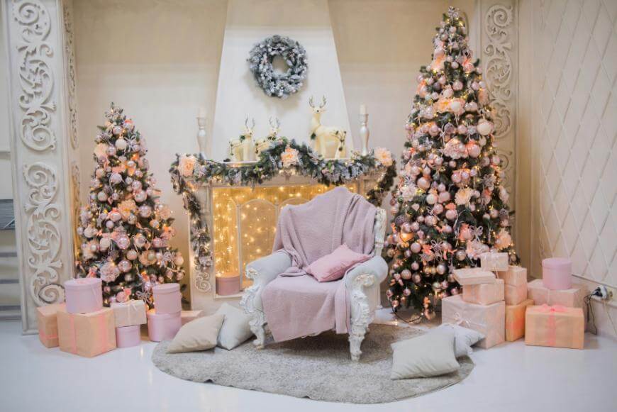 Elegant Christmas scene with two decorated trees, a white ornate armchair draped in pink blankets, pastel gift boxes, pillows on a rug, and a garlanded mantel with lights and reindeer figurines.