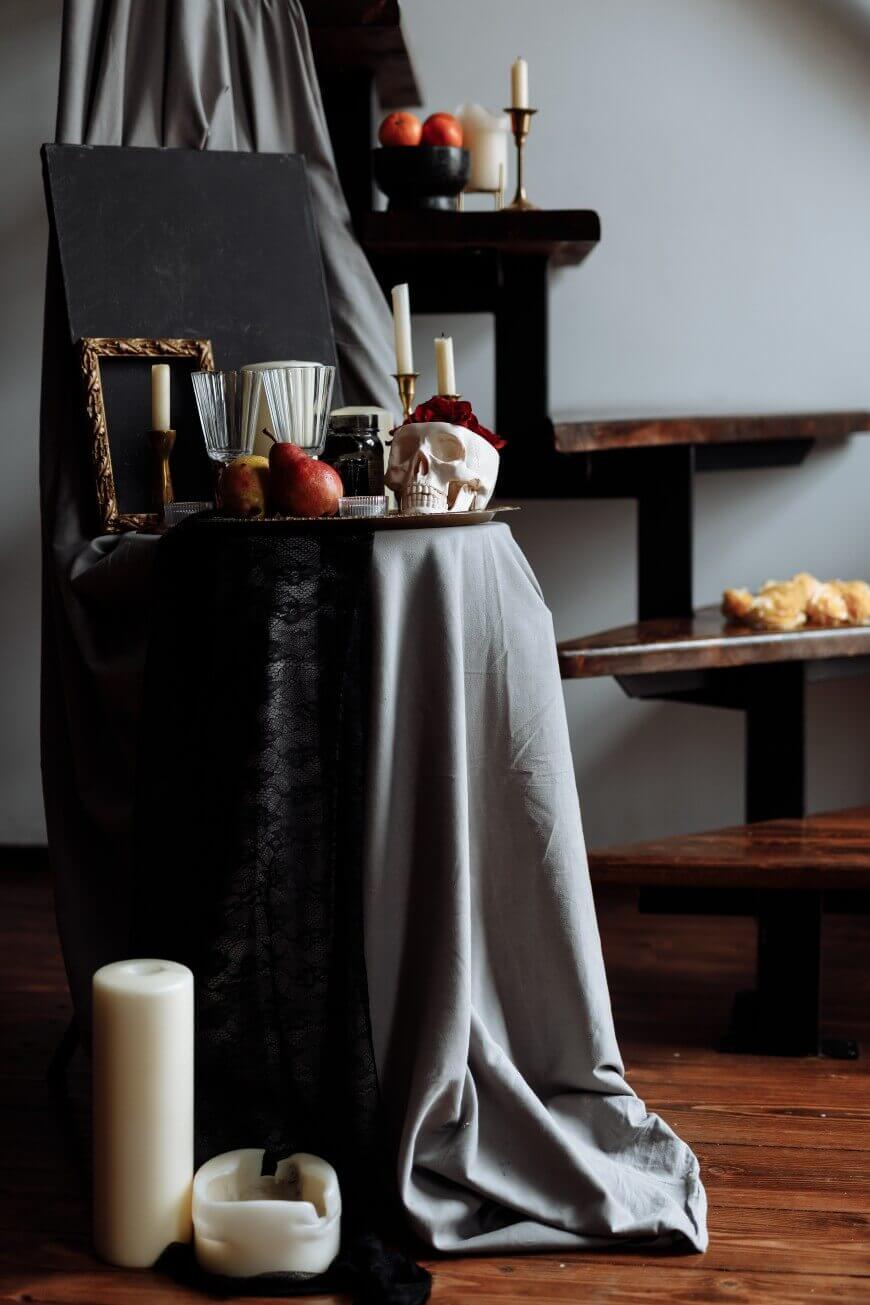 A still life scene on wooden stairs features candles, glasses, apples, a skull decoration, and draped gray and black fabric, with a chalkboard and a gold frame in the background. Croissants sit on a stair step.