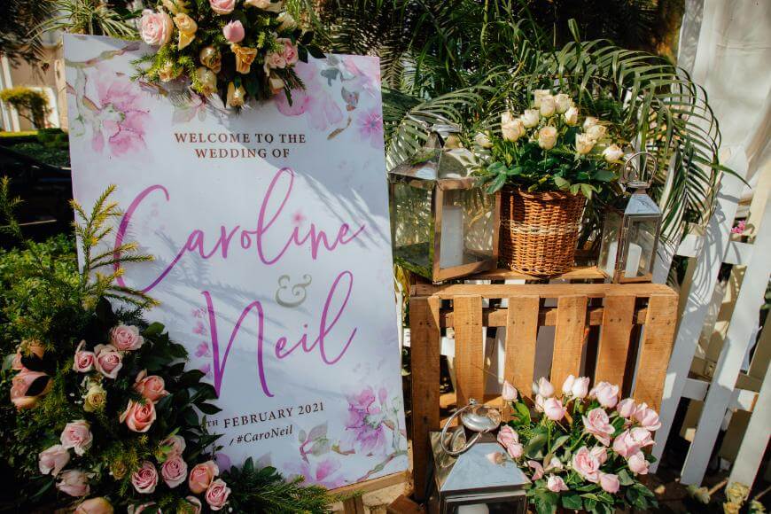 A floral wedding sign reads Welcome to the wedding of Caroline & Neil, 14th February 2021. Surrounded by pink and white roses, greenery, lanterns, and a wicker basket on a wooden pallet.