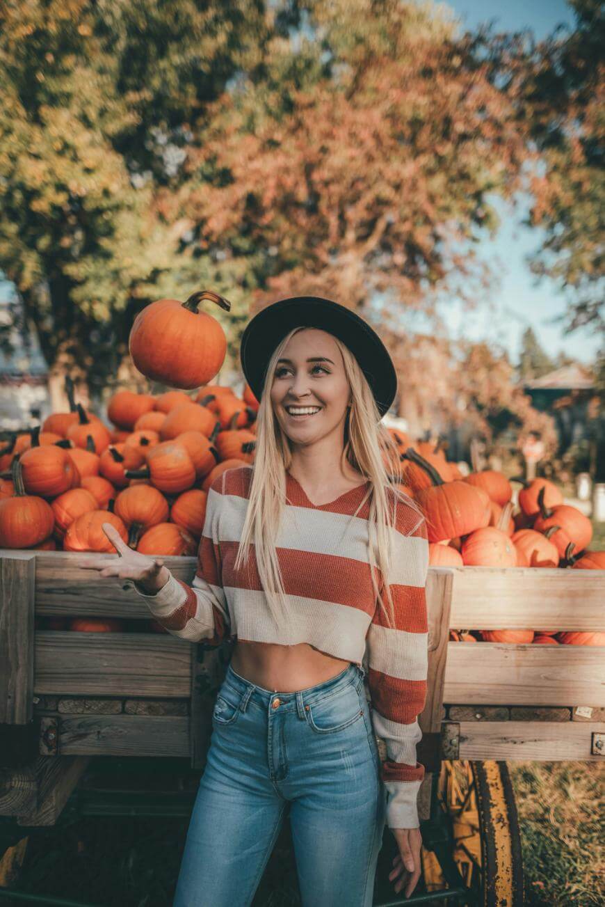 A young woman in a striped crop top and hat stands in front of a wagon full of pumpkins, smiling as she tosses a small pumpkin in the air. Trees with autumn foliage are in the background.