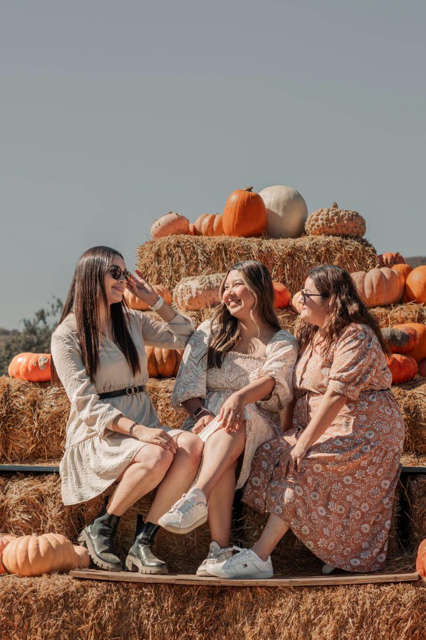 Three women in light-colored dresses sit and laugh together on hay bales decorated with various pumpkins, enjoying a sunny day outdoors against a clear sky.