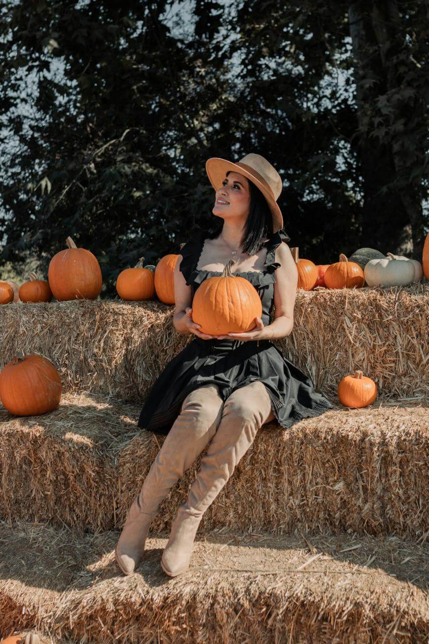 A woman in a black dress, tan hat, and knee-high boots sits on hay bales surrounded by pumpkins, holding one in her lap and looking up, outdoors on a sunny autumn day.