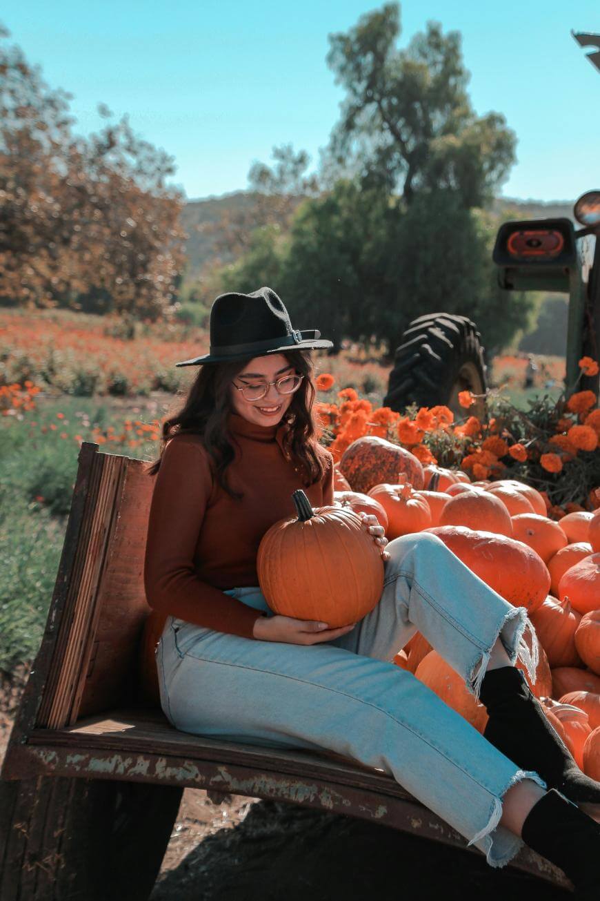 A young woman in a hat and glasses sits on a wooden bench, smiling as she holds a pumpkin. She is surrounded by pumpkins and orange flowers, with a tractor and field in the background on a sunny day.
