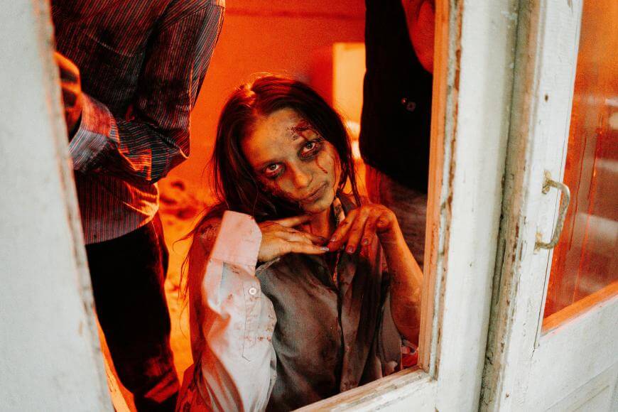 A person with messy hair, pale skin, and dramatic dark makeup resembling a zombie poses eerily behind a window, with two other people partially visible beside them in a dimly lit, orange-tinted room.