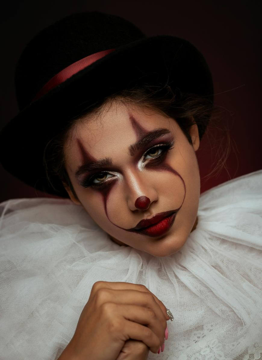 A person dressed as a clown with dramatic makeup, a painted red nose, dark lipstick, a ruffled white collar, and a black hat with a red ribbon, gazes directly at the camera against a dark background.