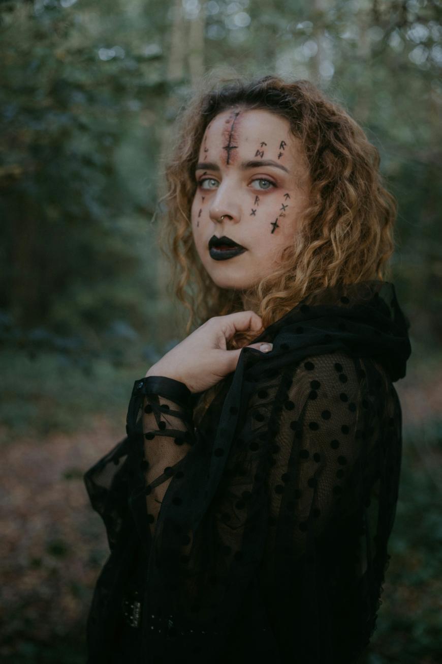 A woman with curly hair and dramatic black makeup, including face paint with crosses and lines, wears a sheer black polka-dot garment and stands in a forest, looking intensely at the camera.