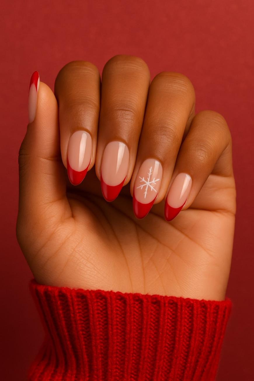 A hand with almond-shaped nails featuring a nude base, red French tips, and one nail decorated with a white snowflake design. The person is wearing a red sweater, and the background is also red.