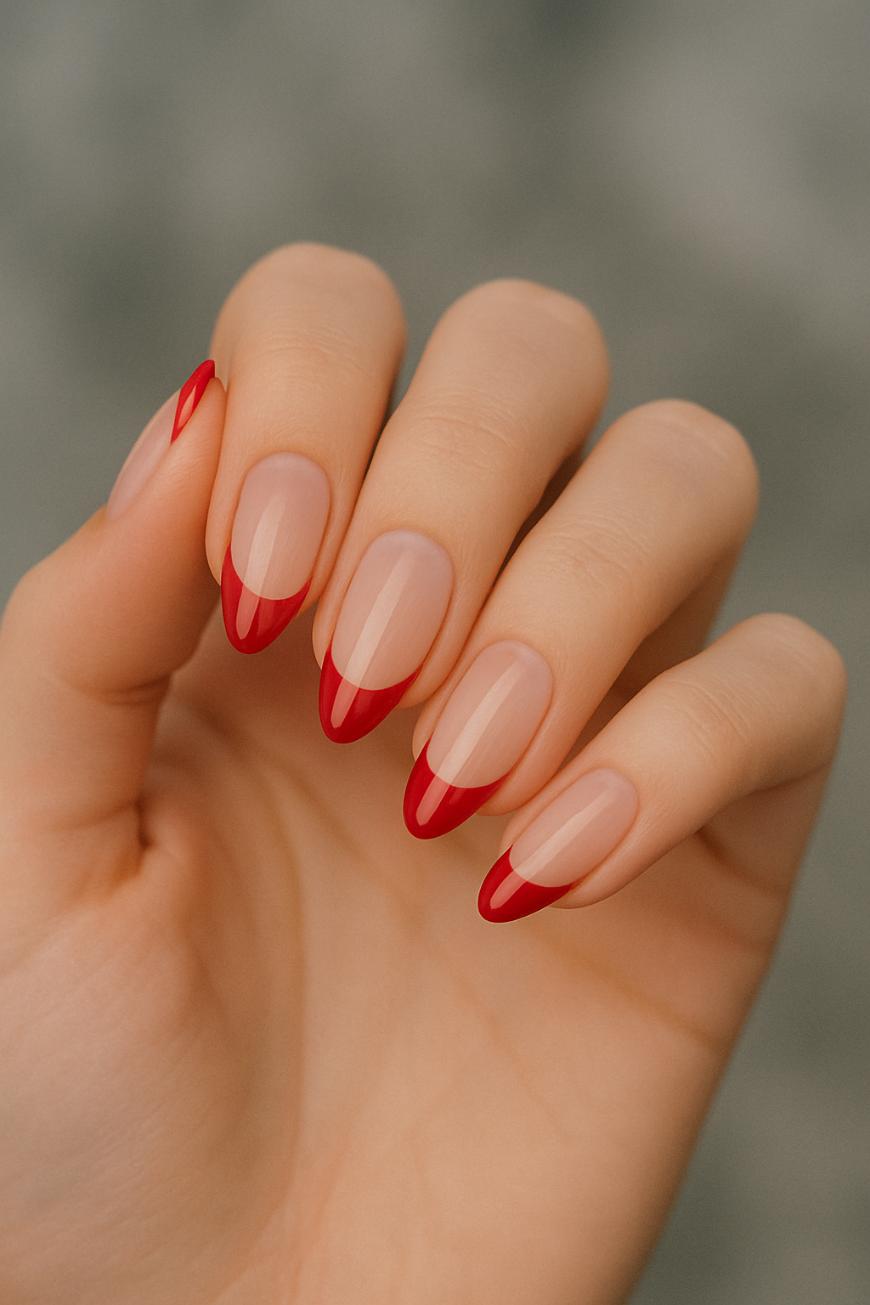 A hand with neatly manicured nails featuring a nude base color and thin, red, pointed French tips. The nails are almond-shaped and the background is blurred gray.