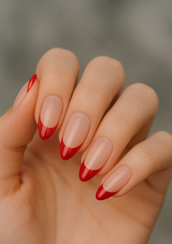 A hand with almond-shaped nails featuring a nude base and glossy red tips in a French manicure style, held against a blurred gray background.