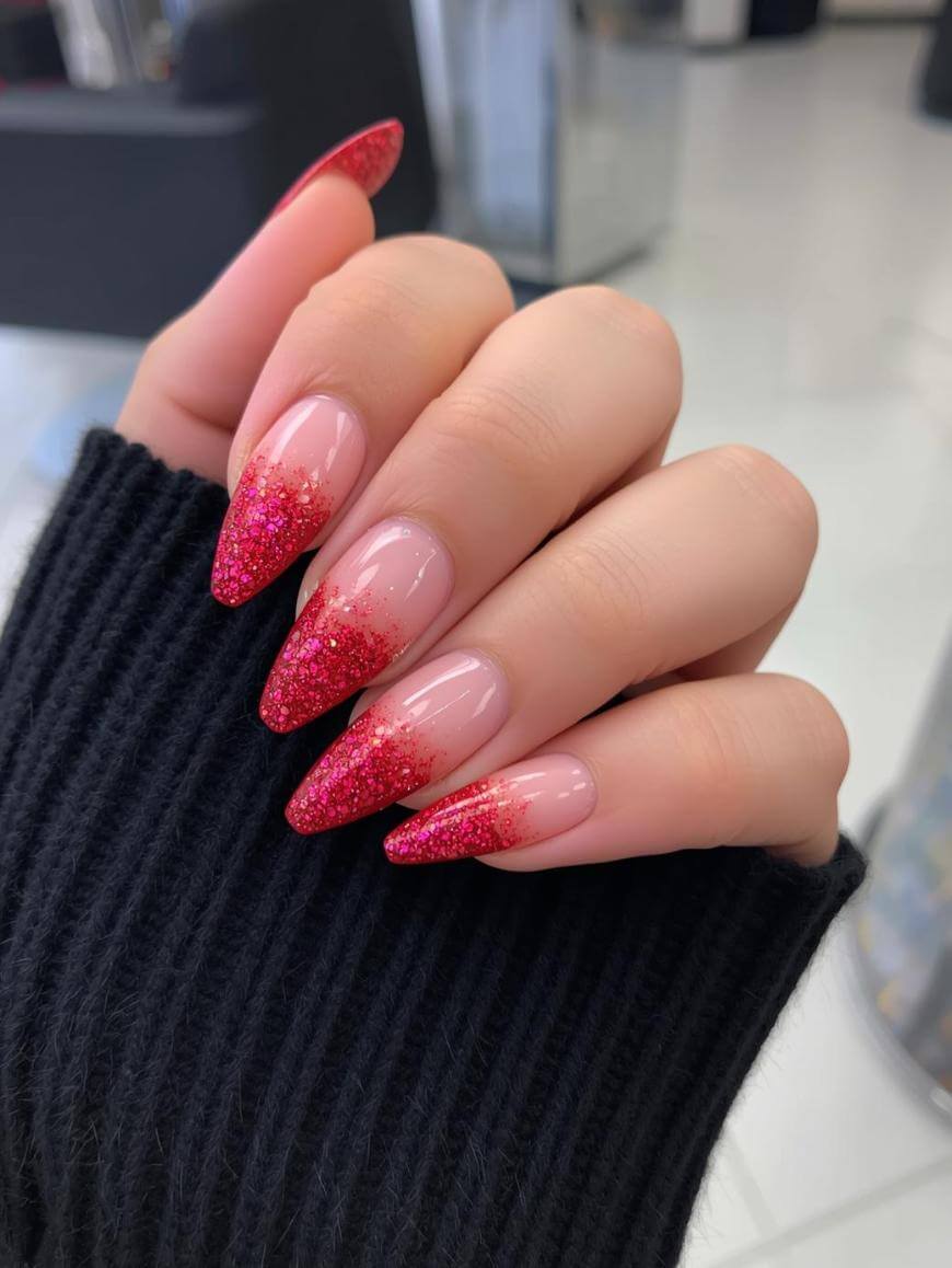A hand with long, almond-shaped nails features a nude base and red glitter ombre tips. The person is wearing a black ribbed sweater, and the background appears to be a modern, bright indoor space.