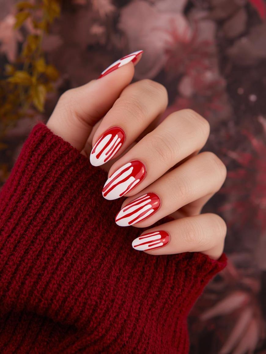 A hand with long, almond-shaped red nails featuring white drip designs, partially covered by a dark red knit sweater, against a blurred floral background.