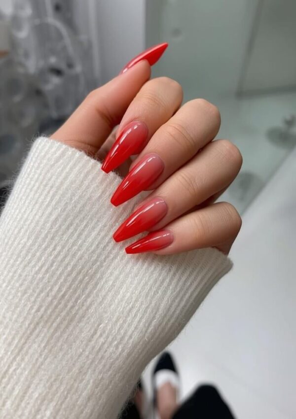 A hand with long, almond-shaped nails painted in a glossy red ombre design is shown against a white background. The person is wearing a cream-colored knit sweater.