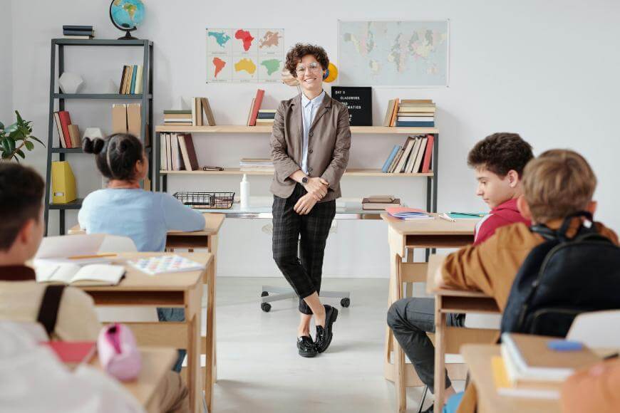A teacher stands smiling at the front of a classroom, facing seated students. Bookshelves, maps, and a globe are visible in the background, creating a welcoming and educational environment.