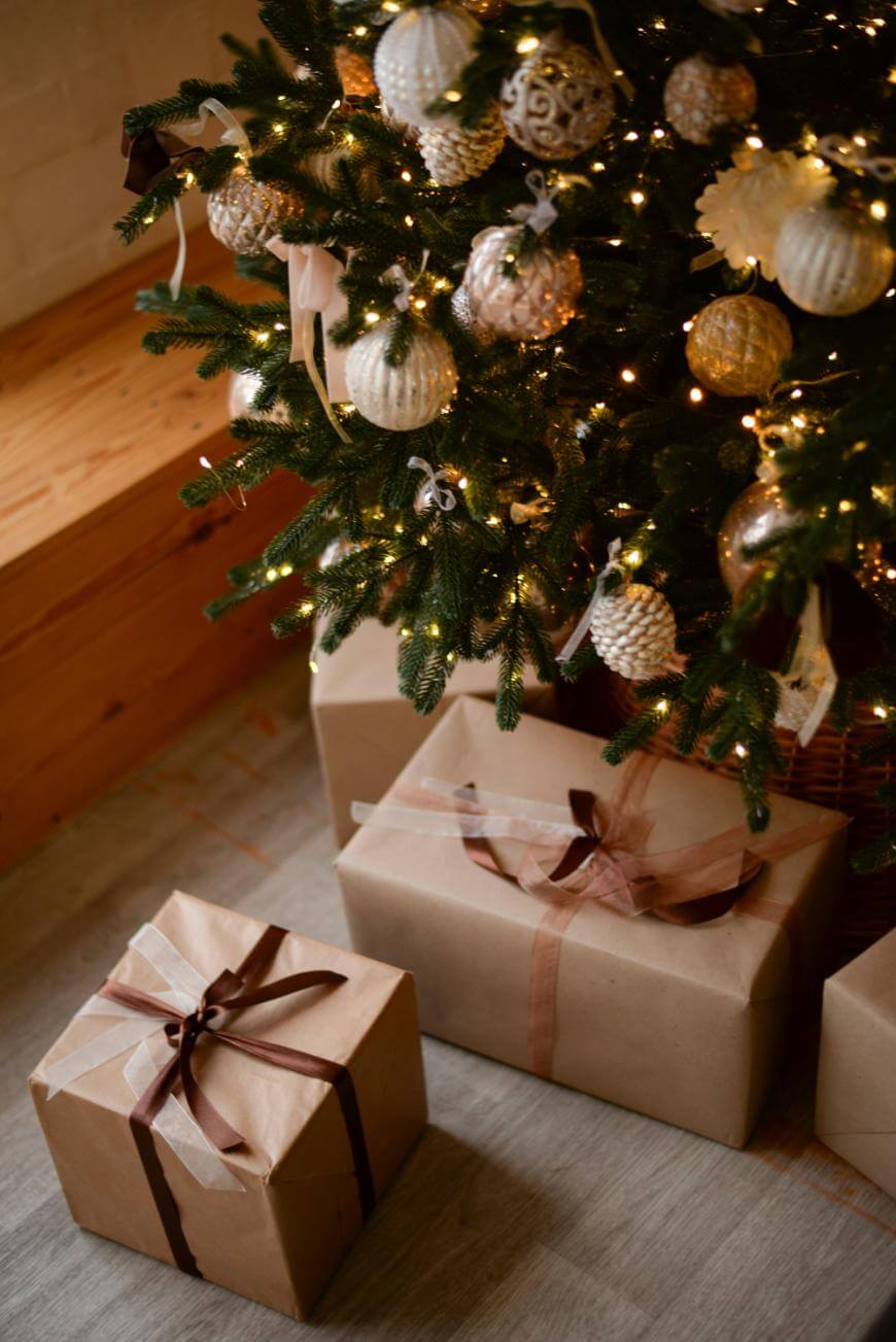 A decorated Christmas tree with gold and white ornaments and string lights stands above several wrapped gifts with ribbon bows on a wooden floor.