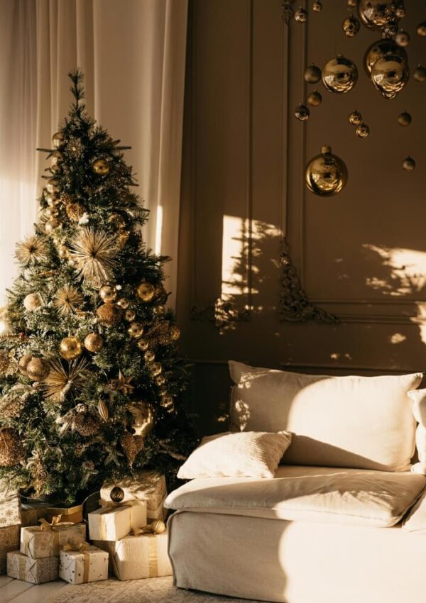 A decorated Christmas tree with gold ornaments and wrapped gifts stands next to a beige sofa in a sunlit, elegant living room with gold baubles hanging from the ceiling.