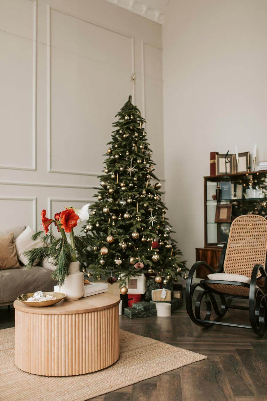 A decorated Christmas tree with gold ornaments stands in a cozy living room with a sofa, a rocking chair, a coffee table with a festive vase, and wrapped gifts under the tree.