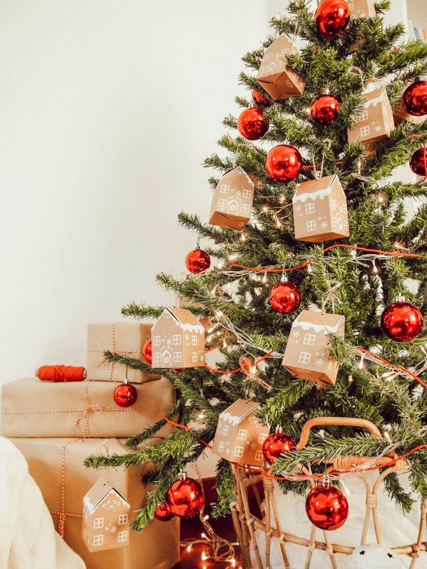 A decorated Christmas tree with red baubles and small house-shaped ornaments stands beside wrapped gifts in brown paper. Warm string lights are intertwined around the tree and presents.