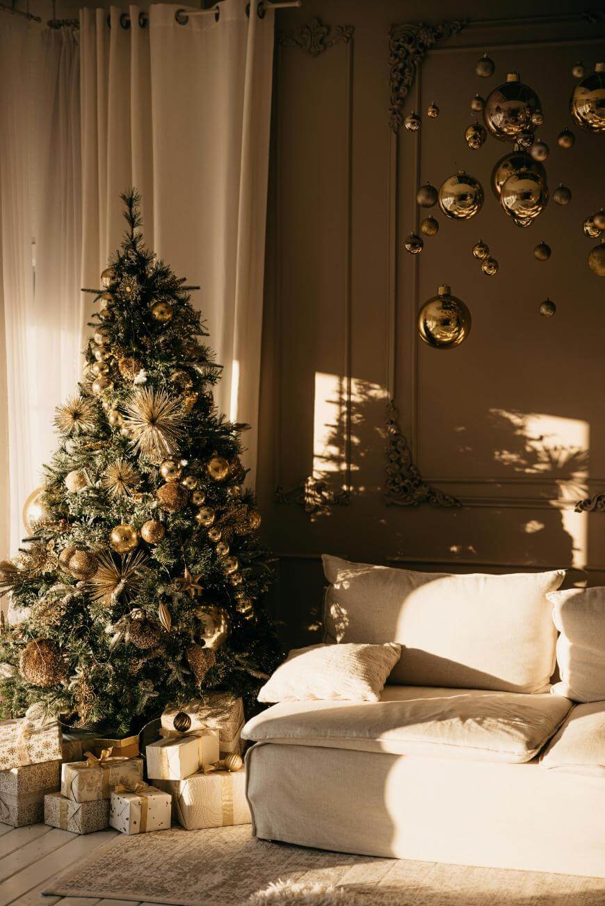 A decorated Christmas tree with gold ornaments and wrapped gifts sits beside a cream-colored sofa in a sunlit, elegant living room with gold wall decorations and sheer curtains.