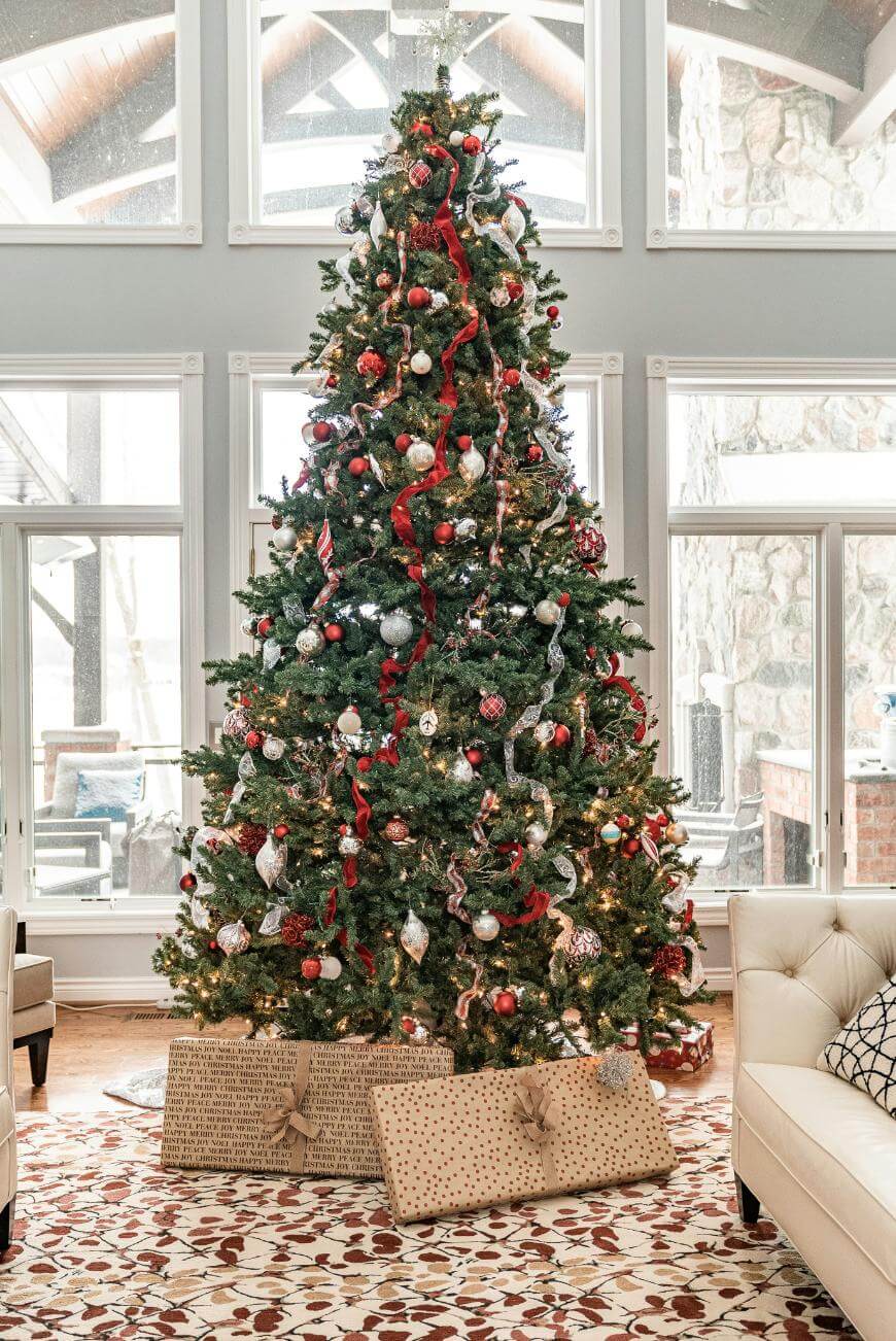 A decorated Christmas tree with red and white ornaments and ribbon stands in front of tall windows, with wrapped presents placed underneath. The room has a patterned rug and beige sofas. Snow is visible outside the windows.