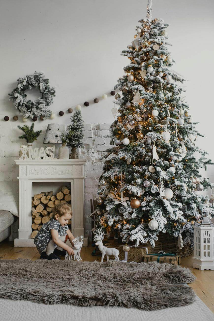 A young child plays with white reindeer figurines near a decorated Christmas tree and a fireplace with stacked logs, festive ornaments, and a wreath in a cozy living room.