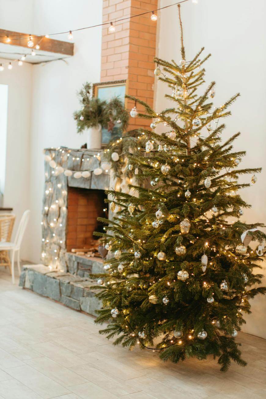 A decorated Christmas tree with silver ornaments and string lights stands near a stone fireplace adorned with festive garland and fairy lights in a cozy, well-lit room.