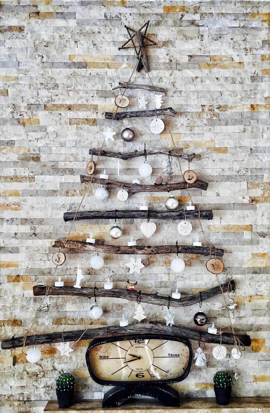 A minimalist Christmas tree made of horizontal wooden branches is decorated with white and silver ornaments and hangs against a stone wall. At the base is a vintage scale and two small potted cacti.