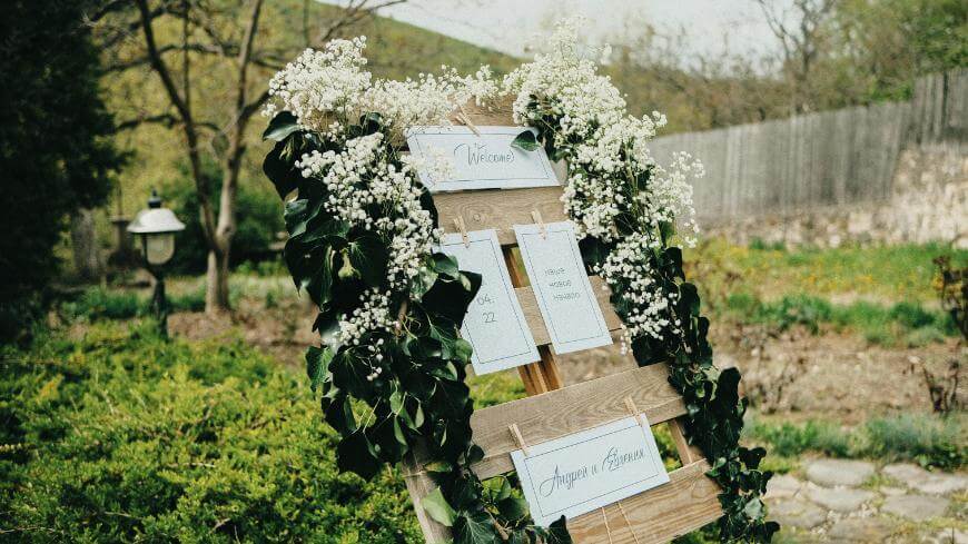 A rustic wooden welcome sign decorated with white flowers and greenery stands outdoors in a garden, displaying paper cards for what appears to be a wedding or event seating chart.