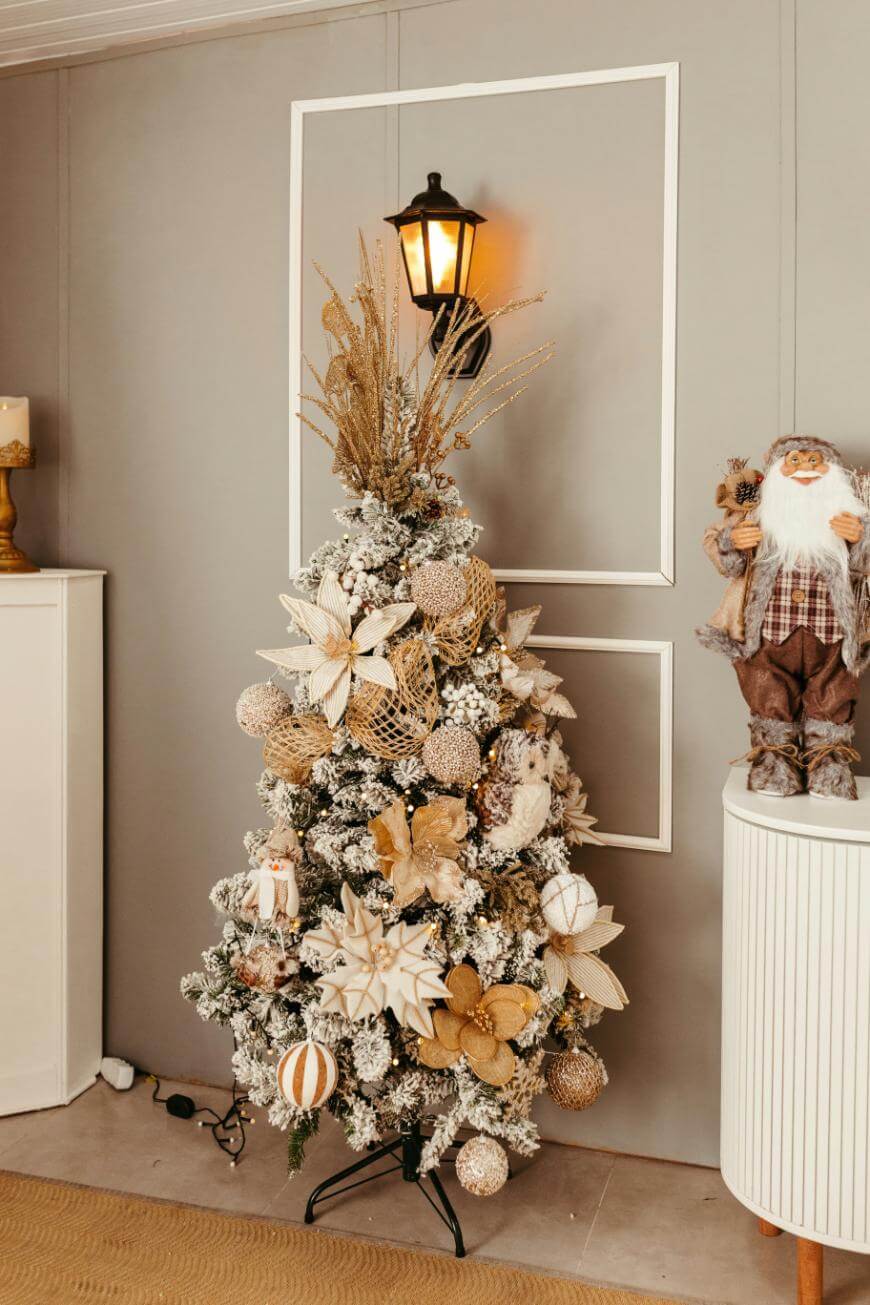 A Christmas tree decorated with white, gold, and beige ornaments and flowers stands by a wall with a lit wall sconce above it. A Santa figurine is displayed on a white cabinet to the right.
