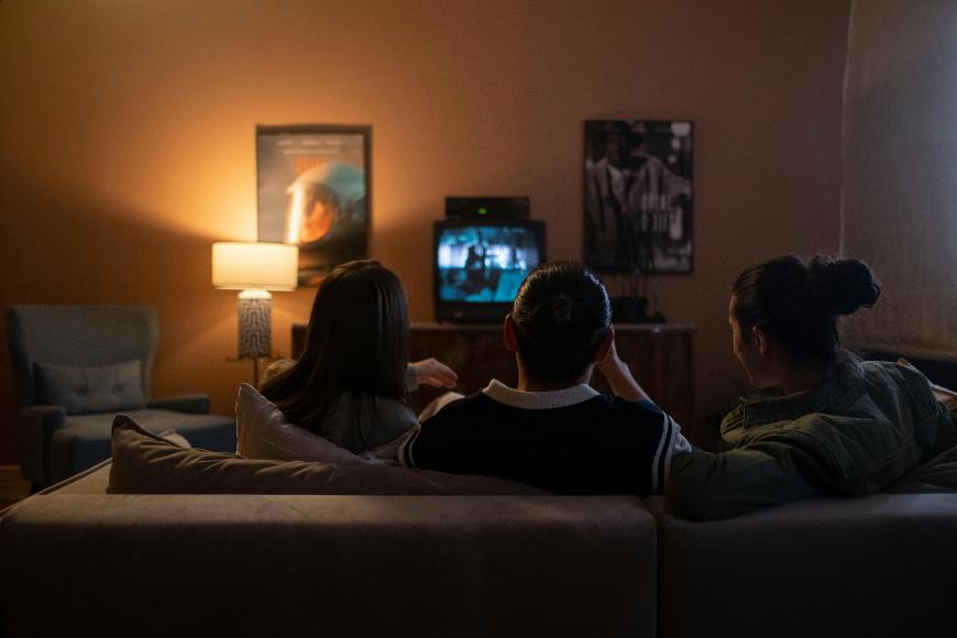 Three people sit on a couch in a dimly lit living room, watching a small TV. Movie posters hang on the walls, and a lit lamp stands next to an empty chair in the background.