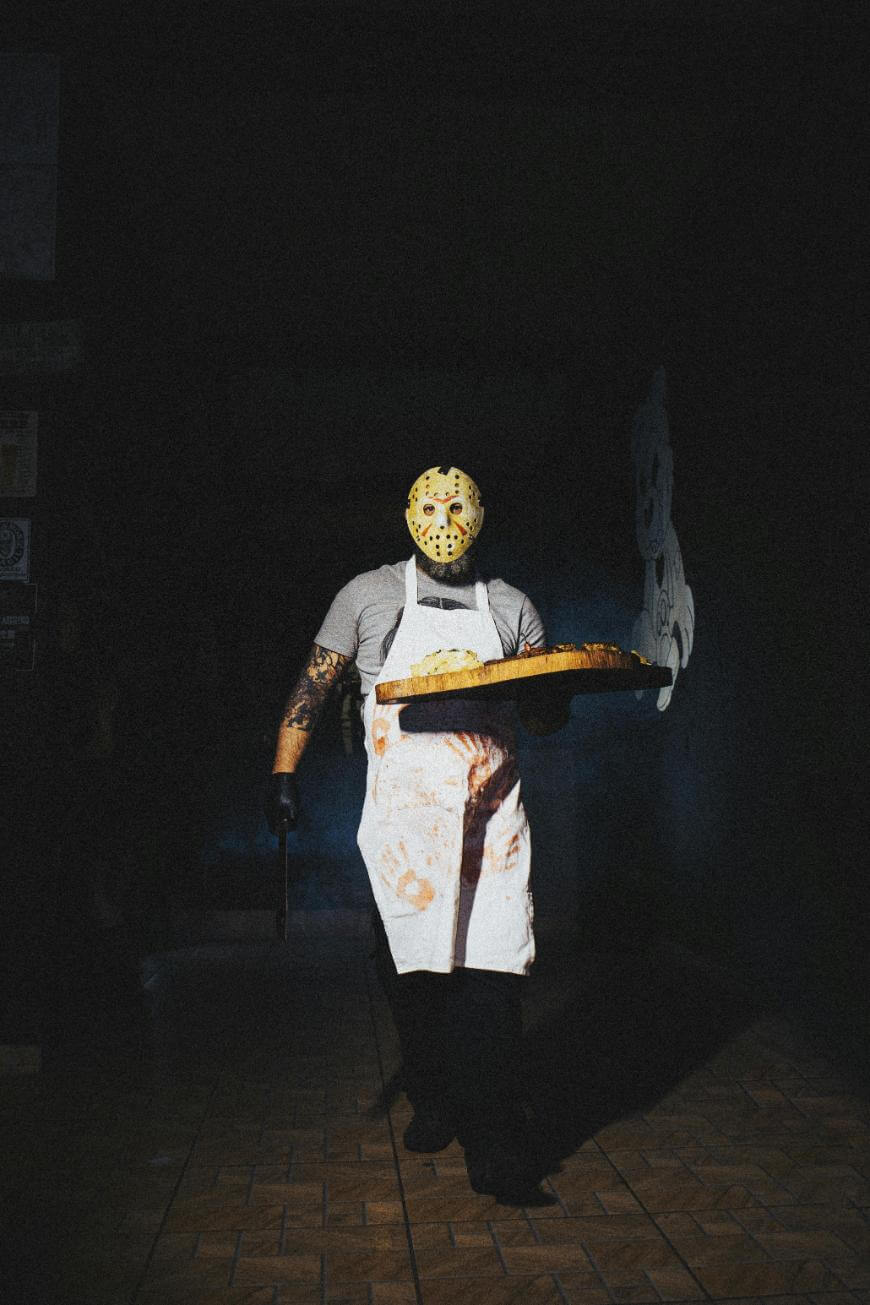 A person in a hockey mask and blood-stained apron holds a large tray of food and a knife, standing in a dimly lit room with a tiled floor.