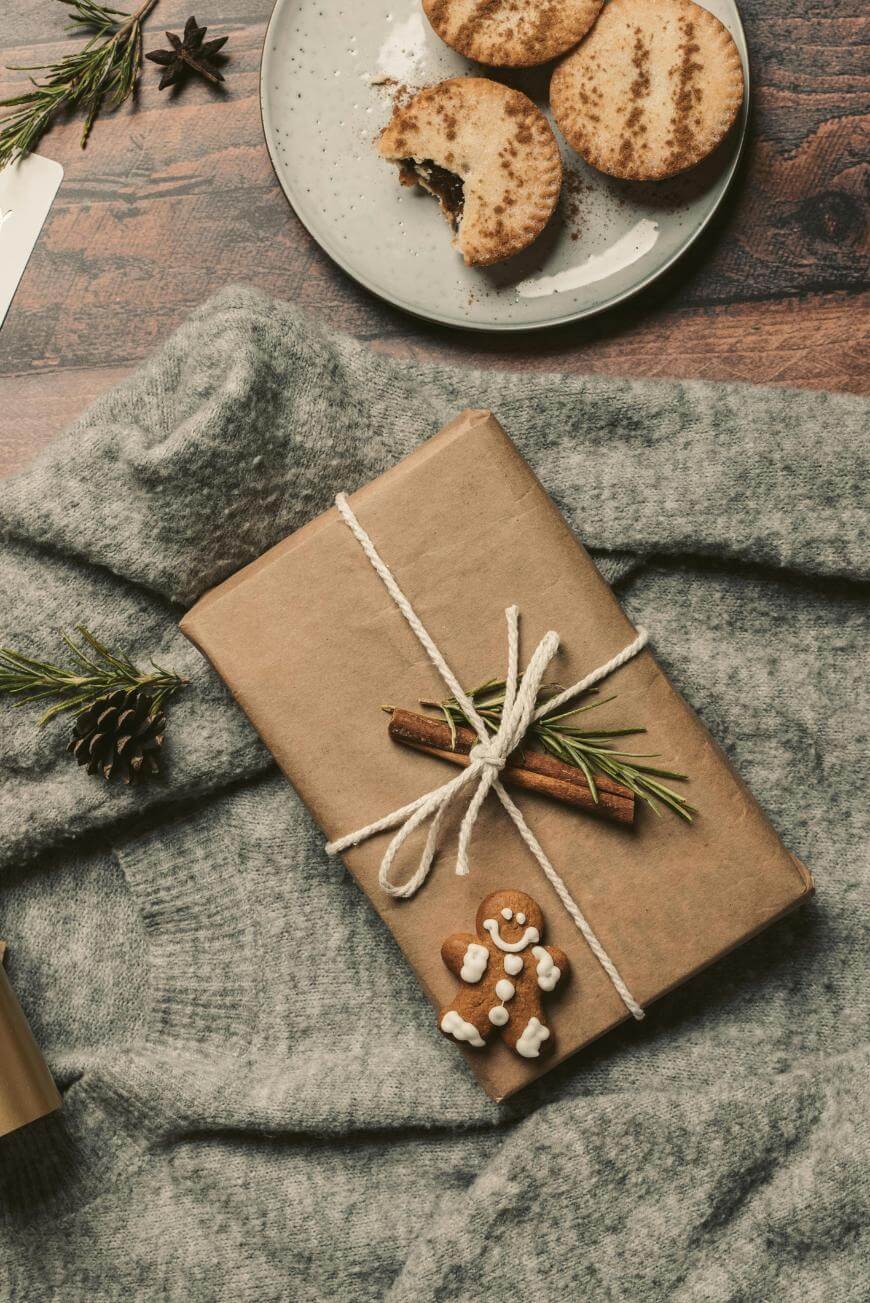A gift wrapped in brown paper with twine, a cinnamon stick, greenery, and a gingerbread man cookie on a gray sweater, with cookies on a plate in the corner on a wooden surface.