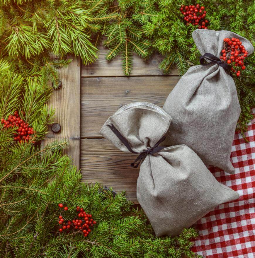 Two linen gift bags tied with black ribbon sit on a wooden surface surrounded by pine branches, red berries, and a red-and-white checkered cloth, creating a festive holiday scene.