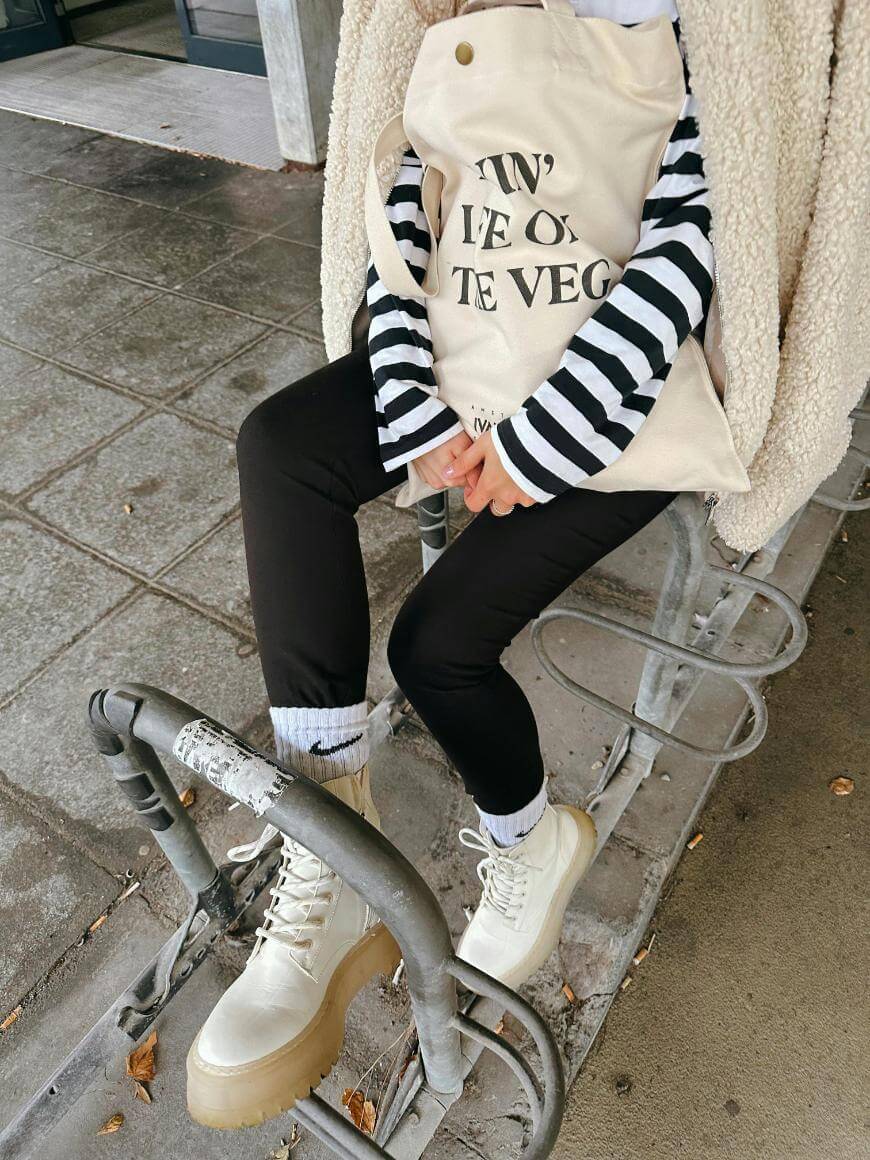 A person wearing white boots, black pants, a striped shirt, and a beige fleece jacket sits on a metal bike rack holding a cream tote bag with black text. The ground is wet and paved with square tiles.