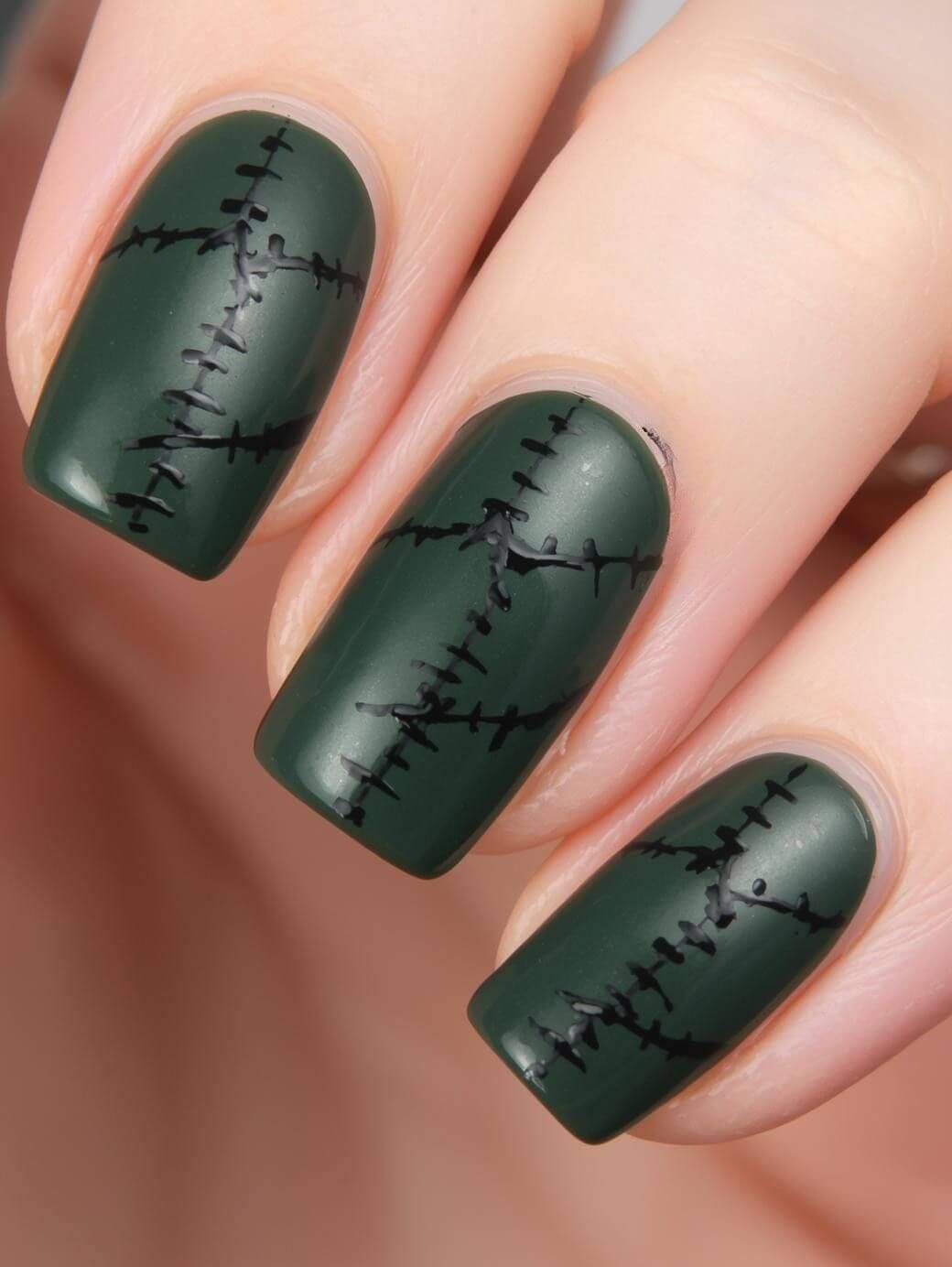A close-up of fingernails painted matte dark green with black stitched line designs, resembling Frankenstein’s monster stitches, on each nail.