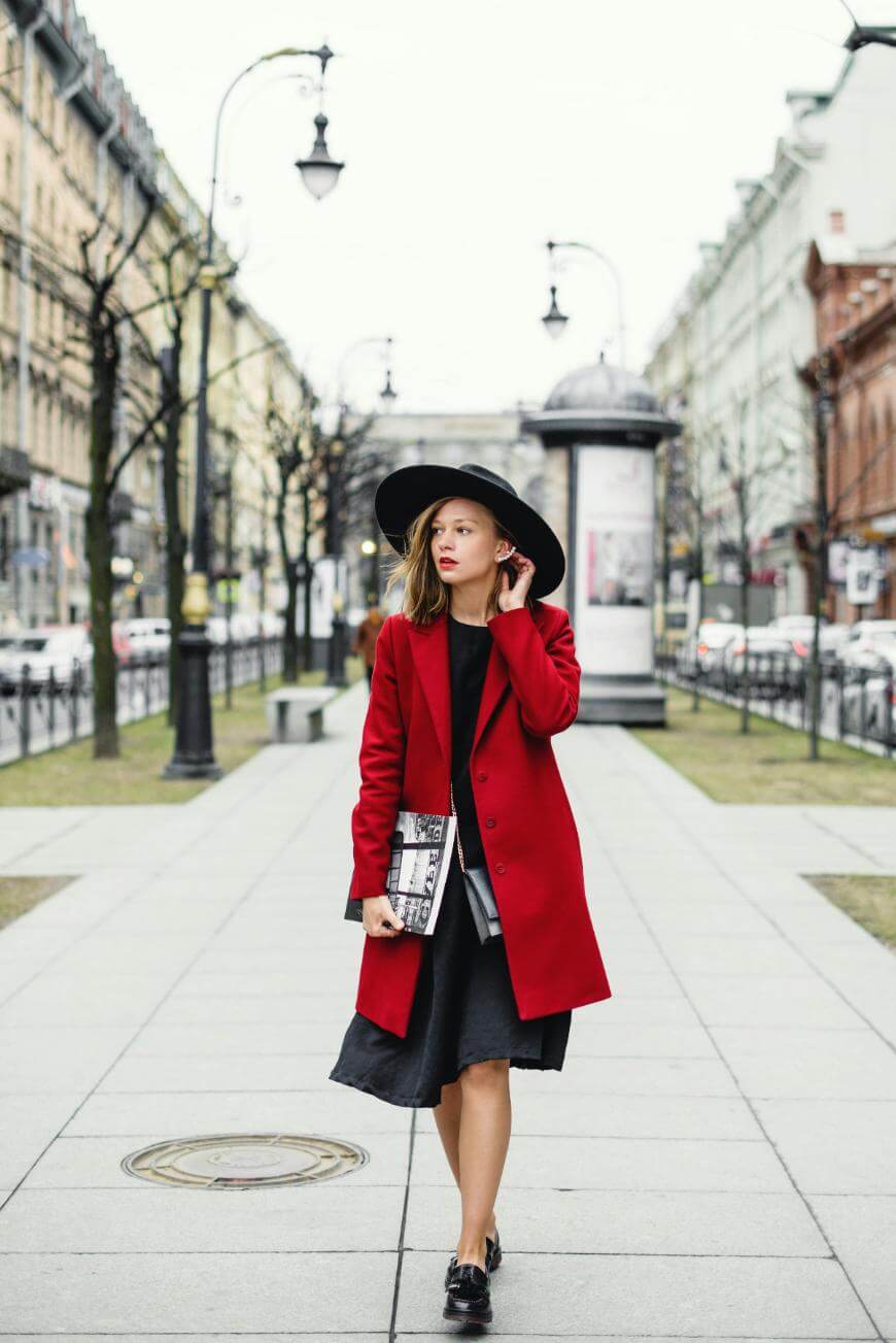 A woman wearing a wide-brimmed black hat, red coat, and black dress walks down a city sidewalk holding a newspaper-print clutch. Leafless trees and buildings line the street on a cloudy day.