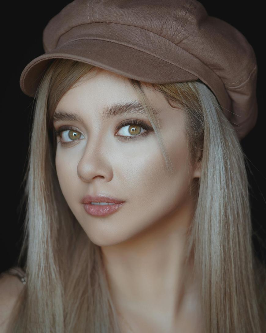 A young woman with long, straight blonde hair wears a brown cap and subtle makeup. She looks directly at the camera with hazel eyes, set against a dark background.