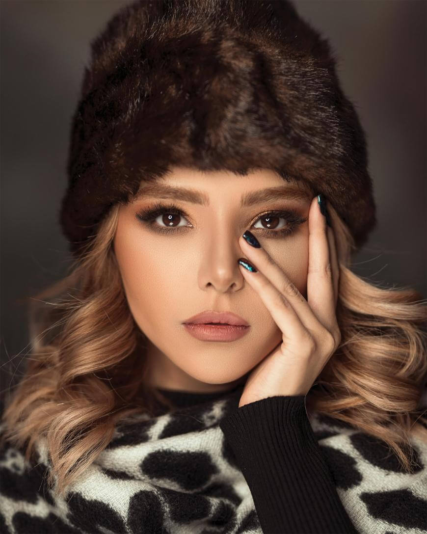 A woman with wavy blonde hair wears a dark fur hat and animal print clothing, gazing at the camera with her hand resting on her face. She has bold eye makeup and manicured nails with black nail polish.