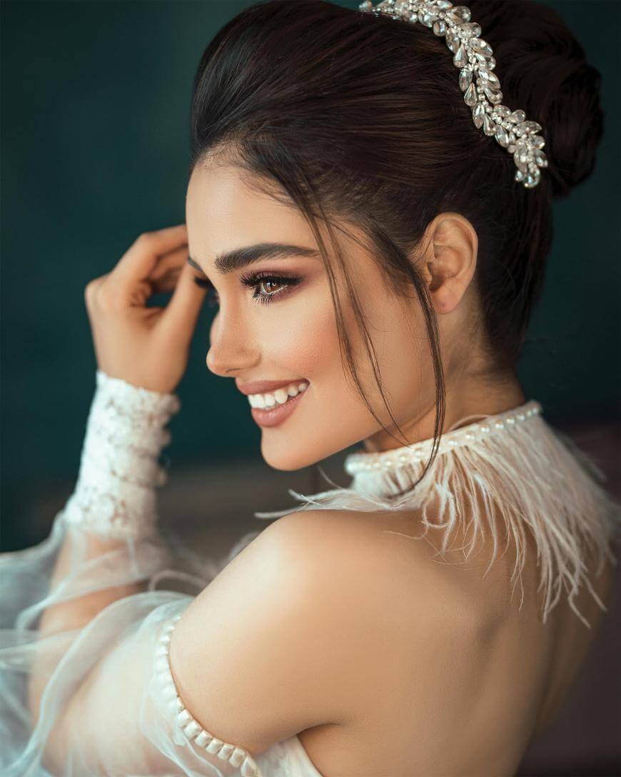 A woman in an elegant off-shoulder white gown with feather details smiles, showing her profile. Her dark hair is styled up with a jeweled hairpiece, and she wears dramatic makeup and long lashes.