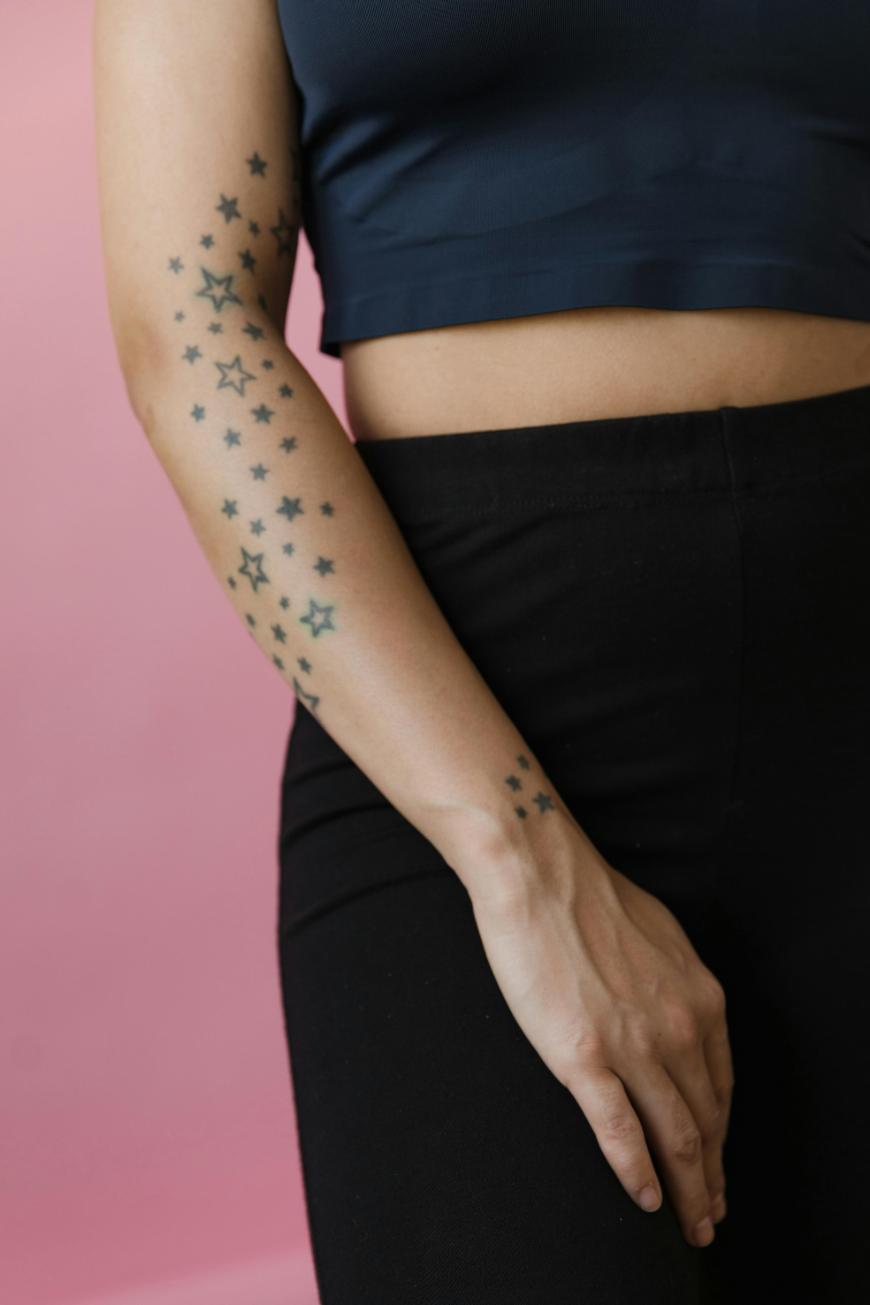 A person standing against a pink background, wearing a dark crop top and black pants, with their arm extended down and covered in various sizes of star tattoos.