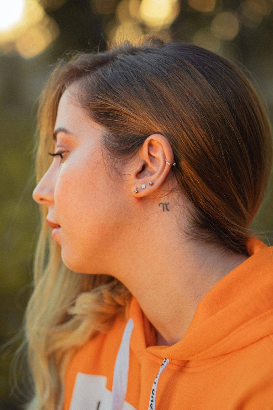 A woman in an orange hoodie is shown in profile, revealing multiple ear piercings and a small pi (π) symbol tattoo on her neck. The background is softly blurred.