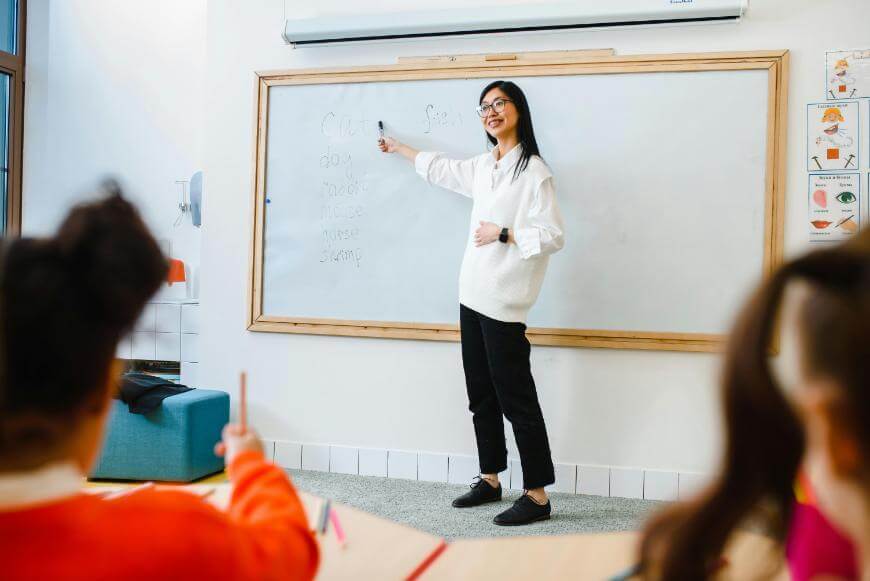 A teacher stands at a whiteboard, smiling and holding a marker while pointing to words written on the board. Two students sit at desks in the foreground, facing the teacher.